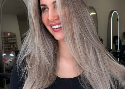 Woman with long, silver-blonde hair smiling in a salon with mirrors in the background.