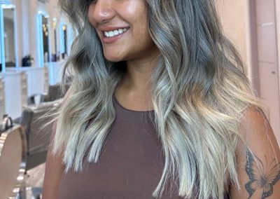 Smiling woman with wavy, ash blonde hair and a bird tattoo on her arm standing in a salon.