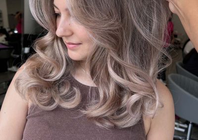 Woman with voluminous, wavy, ash-blonde hair and curtain bangs in a salon, wearing a brown tank top.