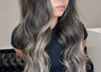 Woman with long, wavy, ash-brown hair with blonde highlights, wearing a black outfit, standing indoors.