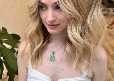 Woman with wavy blonde hair in a white dress, standing indoors; a hand lifts part of her hair.