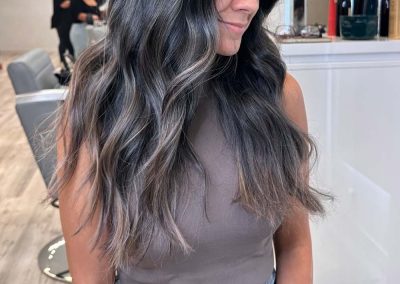 Woman with long, wavy dark hair and gray highlights sitting in a salon, wearing a sleeveless top and jeans.