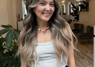 Smiling woman with long, wavy blonde hair stands indoors wearing a gray top and colorful necklace.