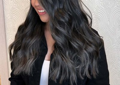 Woman with long, wavy black and gray hair, wearing a black jacket, smiling and looking down.