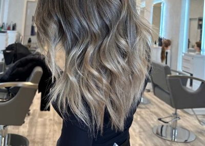 Woman with wavy, shoulder-length ash blonde balayage hair in a modern hair salon.