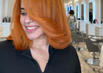 Woman with shoulder-length, vibrant orange hair smiles in a modern, brightly lit hair salon.