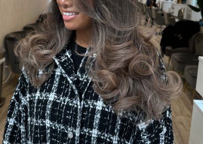 Woman with voluminous, wavy, ash-brown hair smiling in a modern, well-lit hair salon.