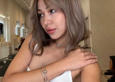 Woman with shoulder-length ash brown hair takes a selfie in a salon, wearing a white tank top and silver jewelry.
