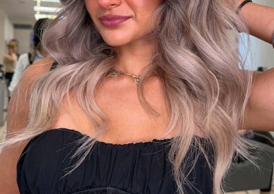 Woman with long, wavy, ash blonde hair wearing a black strapless top, posing in a salon.