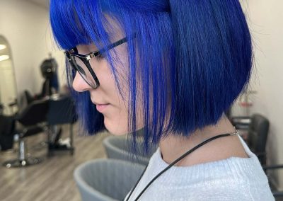 Person with bright blue bob haircut and glasses, wearing a white sweater, in a modern salon.
