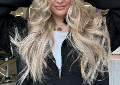 Young woman with long, wavy blonde hair wearing a black zip-up jacket poses indoors with hands in her hair.