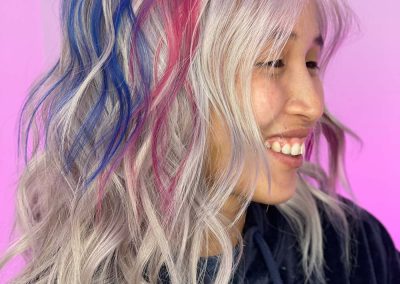 Smiling person with wavy silver hair, featuring blue and pink streaks, wearing a navy hoodie.