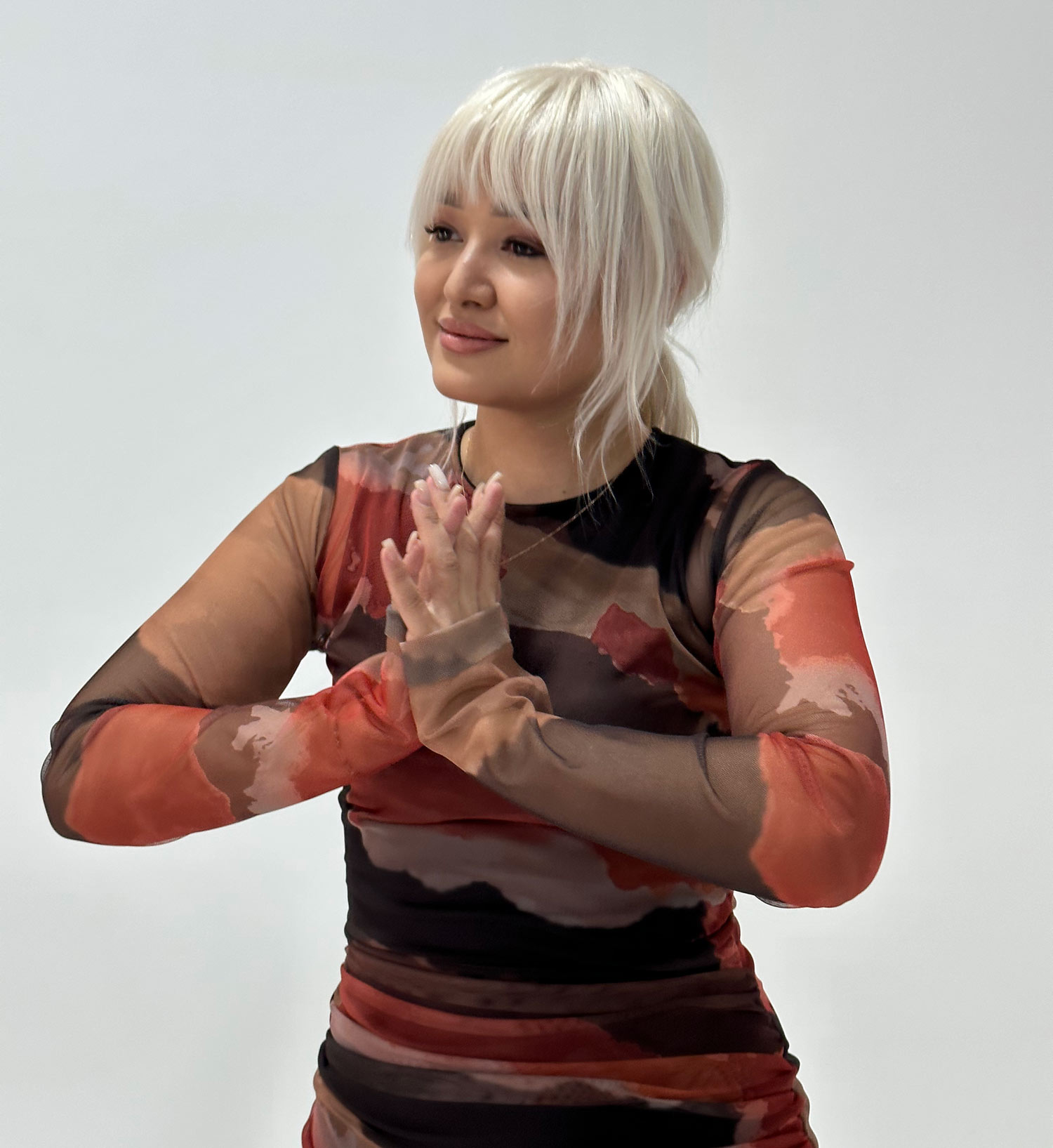 Woman with platinum blonde hair in a patterned long-sleeve dress standing with hands clasped, smiling.