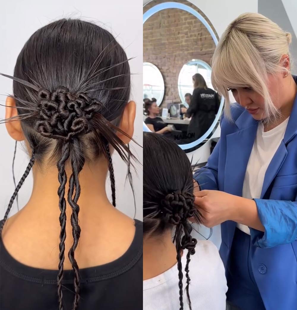 A stylist braids a girl's hair into an intricate bun with loose, braided strands hanging down.