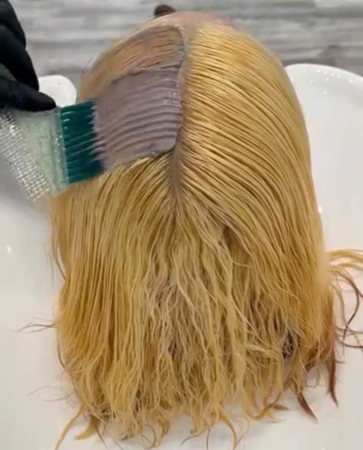A gloved hand applies hair dye to the roots of wet, blond hair with a brush.