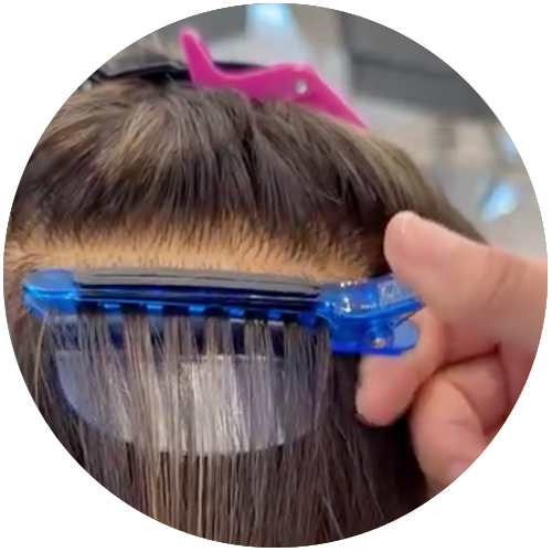 A hand uses a blue hair extension tool to attach extensions to brown hair near the scalp.