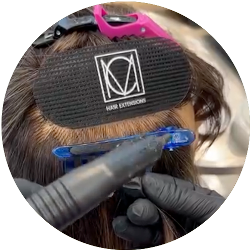 Gloved hands applying hair extensions to dark hair using a sectioning tool and clips.